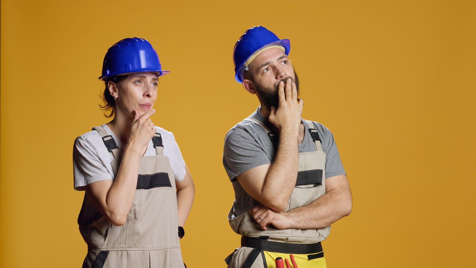 Deux ouvriers du bâtiment portant des casques bleus et des combinaisons de travail se tiennent debout sur un fond jaune, tous deux avec des expressions pensives et les mains sur le menton.
